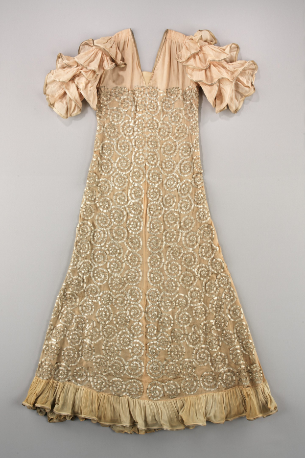 Ein cremefarbenes Kleid mit goldener Stickerei und Rüschen, das auf einer weißen Oberfläche präsentiert wird.