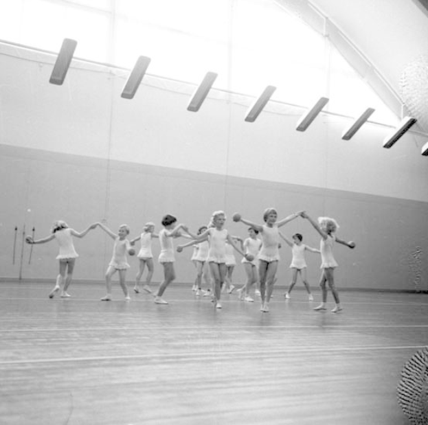 Schwarzes und weißes Bild von jungen Mädchen, die sich in einem Studio mit einem Ball auf der rechten Seite und Lampen an der Decke halten und tanzen.