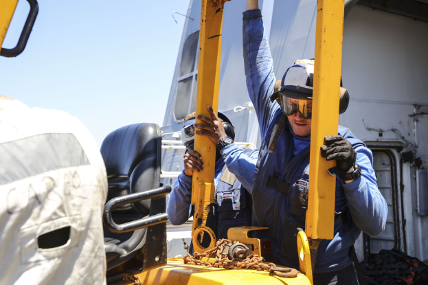 Zwei Männer in blauen Uniformen und Schutzausrüstung arbeiten an einem gelben Gabelstapler, wobei einer ein Metallobjekt hält, in der Nähe eines Fahrzeugs unter einem Himmel.