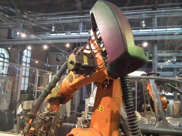 Roboterarm bei der Montage eines Autos in einer industriellen Fabrikumgebung mit Metallstangen, Beleuchtung und Hintergrundmaschinerie.