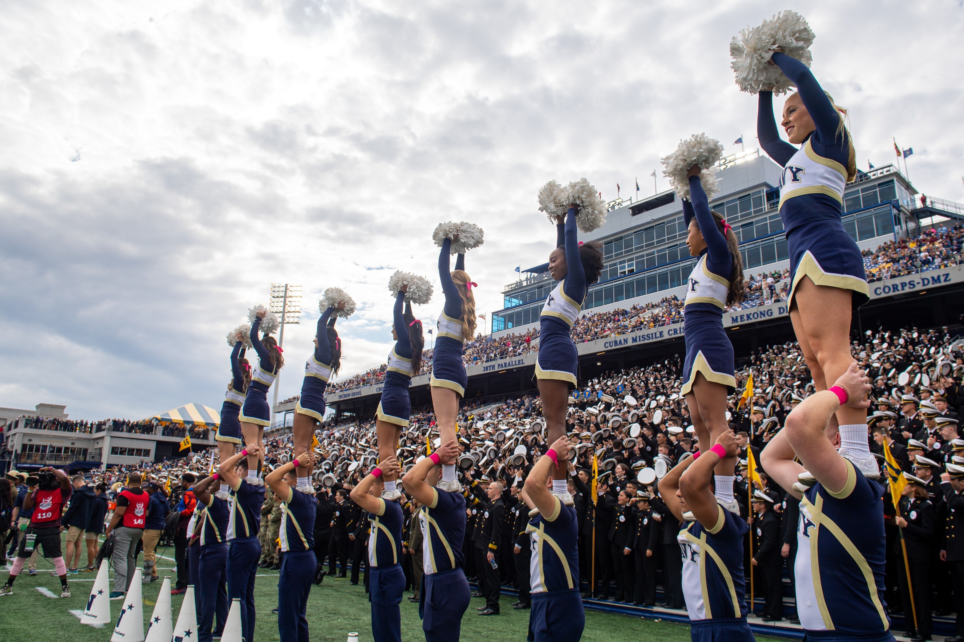 Eine Gruppe von Cheerleadern in blauen und weißen Uniformen führt einen Stunt aufeinander aus, während sie Pompons halten, mit einer Menge und Stadionstrukturen im Hintergrund.