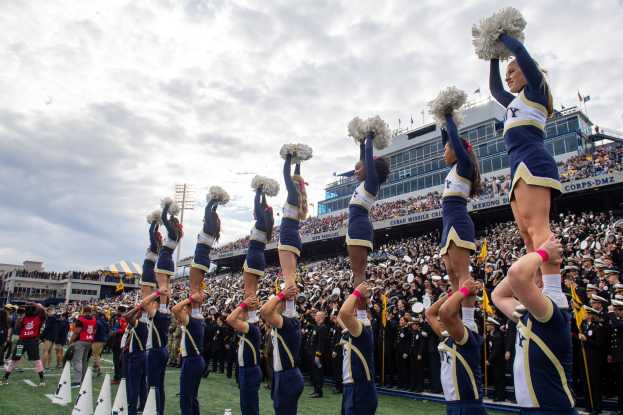 Eine Gruppe von Cheerleadern in blauen und weißen Uniformen führt einen Stunt aufeinander aus, während sie Pompons halten, mit einer Menge und Stadionstrukturen im Hintergrund.
