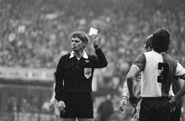 Ein Schiedsrichter in einem Schwarz-Weiß-Bild hält eine rote Karte vor einer Menge von Menschen in Fußballtrikots hoch, wobei ein Zaun im Hintergrund zu sehen ist.