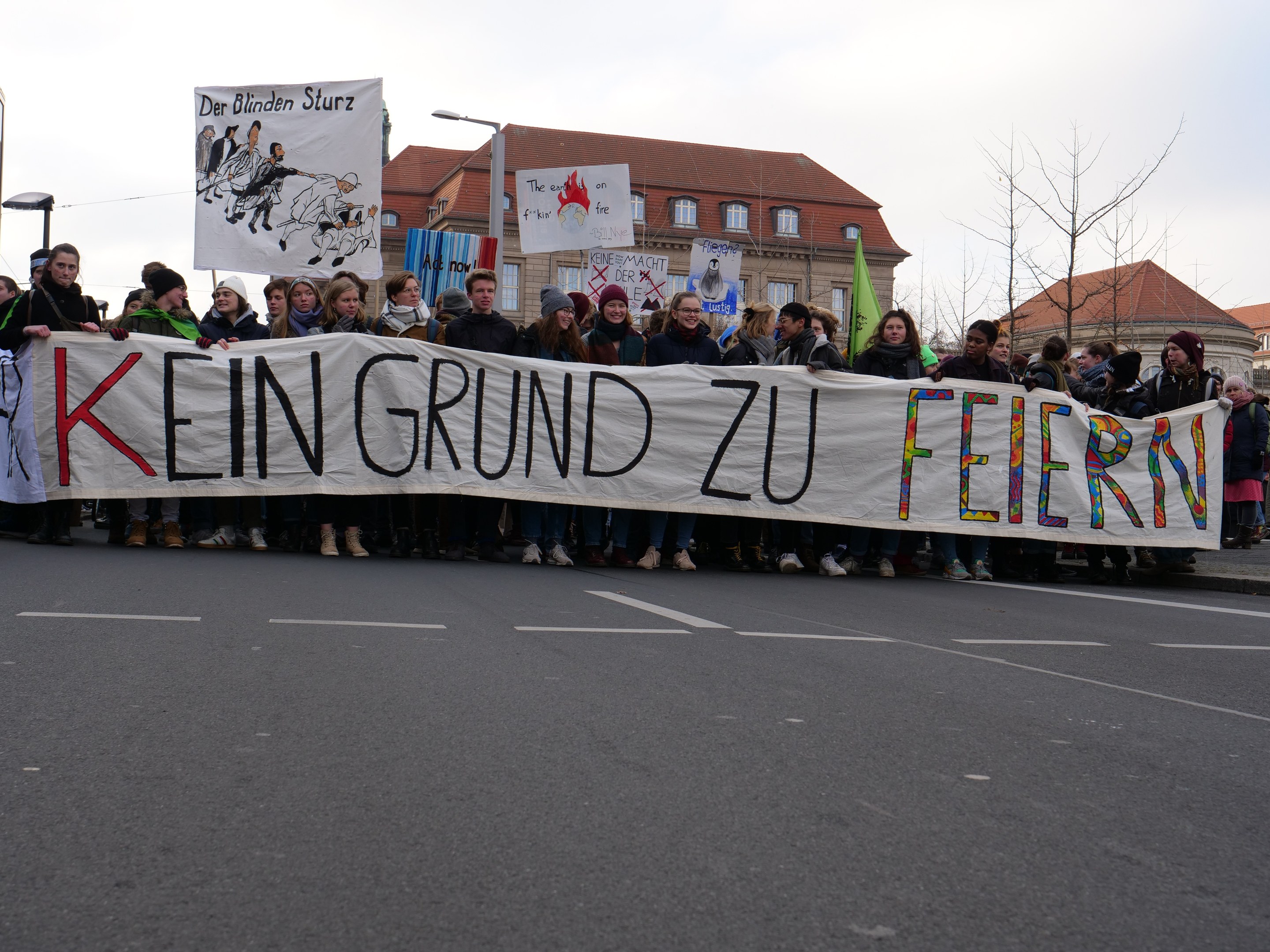 Demonstranten mit einem Transparent mit der Aufschrift 'Kein Grund zu Feiern' gegen deutsche Sparmaßnahmen, mit Straßeninfrastruktur und Gebäuden im Hintergrund.