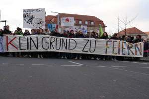 Demonstranten mit einem Transparent mit der Aufschrift 'Kein Grund zu Feiern' gegen deutsche Sparmaßnahmen, mit Straßeninfrastruktur und Gebäuden im Hintergrund.