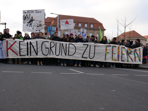 Demonstranten mit einem Transparent mit der Aufschrift 'Kein Grund zu Feiern' gegen deutsche Sparmaßnahmen, mit Straßeninfrastruktur und Gebäuden im Hintergrund.