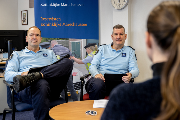 Zwei Polizeibeamte sitzen an einem Tisch mit einer Frau, Papieren und Gegenständen auf dem Tisch, einer Wand mit einem Banner, Uhr und Foto Rahmen im Hintergrund und einem weiteren Tisch mit einem Monitor, Tastatur und Maus links.