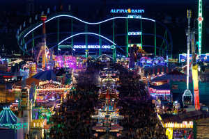 Eine große Menschenmenge versammelt sich um einen hell erleuchteten Karneval in der Nacht, mit Vergnügungsgeräten, Masten, Flaggen und Namensschildern im Hintergrund.