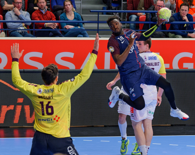 Eine Gruppe von Männern, die auf einem Platz Handball spielen, mit einem Ball in der Luft und Zuschauern im Hintergrund, die das Spiel beobachten, mit einem Schild, auf dem "Futsal-Weltmeisterschaft 2019 - Paris Saint-Germain vs Bordeaux" steht.