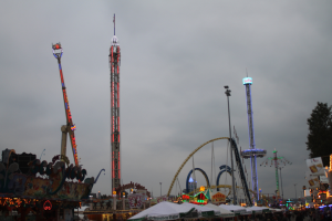Eine Menschenmenge umringt einen hell erleuchteten Faschingsumzug bei Nacht, mit beleuchteten Fahrgeschäften, Pfählen und Bäumen unter einem bewölkten Himmel.
