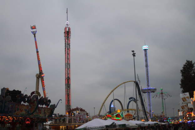 Eine Menschenmenge umringt einen hell erleuchteten Faschingsumzug bei Nacht, mit beleuchteten Fahrgeschäften, Pfählen und Bäumen unter einem bewölkten Himmel.