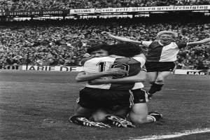 Drei Fußballspieler umarmen sich auf dem Boden und feiern ein Tor vor einer jubelnden Menge in einer schwarz-weißen Stadion-Szene.