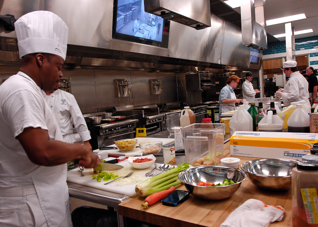 Eine Gruppe von Köchen in einer Küche, die Essen auf einem Schneidbrett zubereiten, mit einem Koch im Vordergrund in einer weißen Schürze und Mütze, der ein Messer hält, und umgeben von Utensilien, Schüsseln, Gemüse, einem Handy, Flaschen und anderen Gegenständen, mit einem Fernseher und Deckenleuchten im Hintergrund.