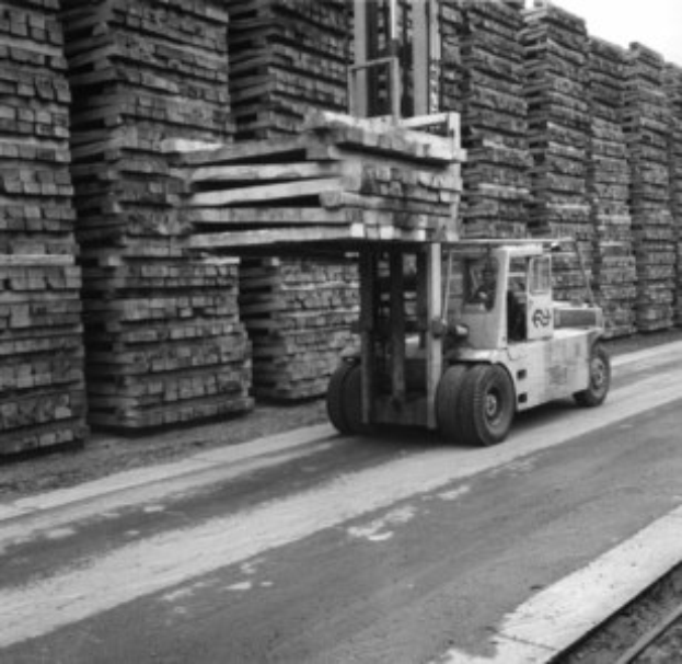 Schwarz-weißes Foto eines Gabelstaplers mit Fahrer auf einer Straße neben einer Bahnschiene, mit gestapelten Holzplanken im Hintergrund.