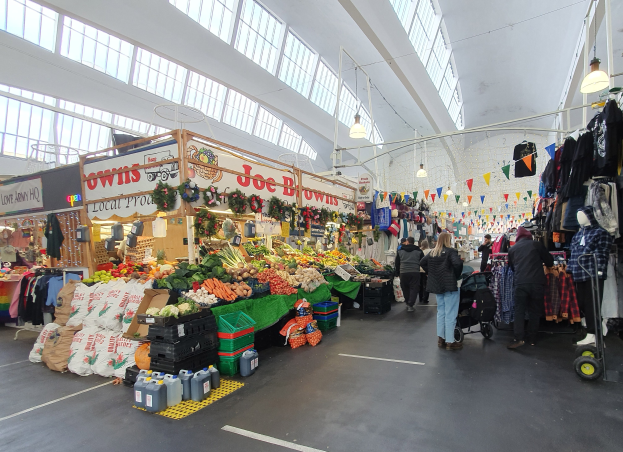 Ein belebter Indoor-Markt mit Menschen, die zwischen Ständen mit Obst, Gemüse und hängenden Kleidern entlanggehen, unter Deckenlampen mit großen Glasfenstern im Hintergrund.