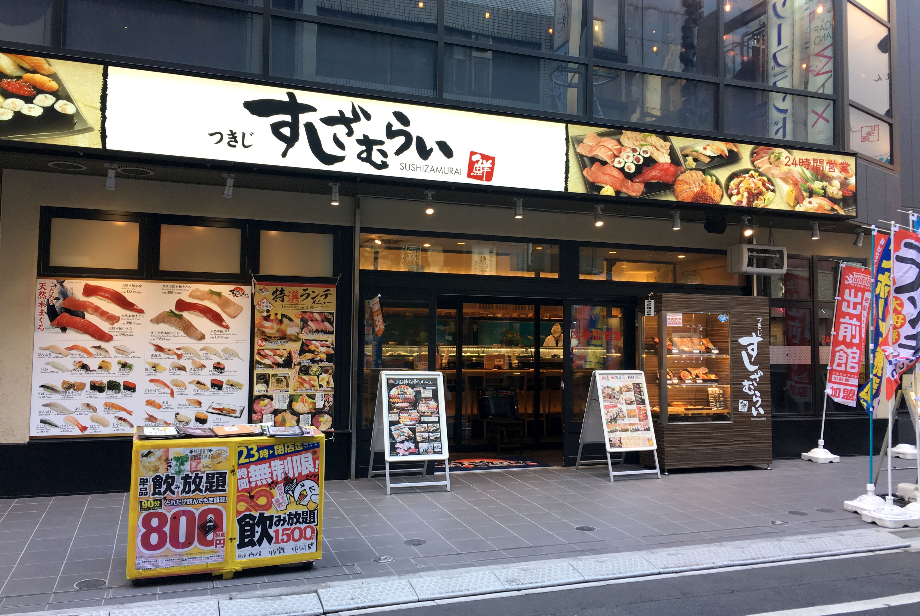 Außenansicht einer Sushi-Bar mit Glaswänden, Essensauslagen und Schildern an einer Straßenecke in Tokyo.