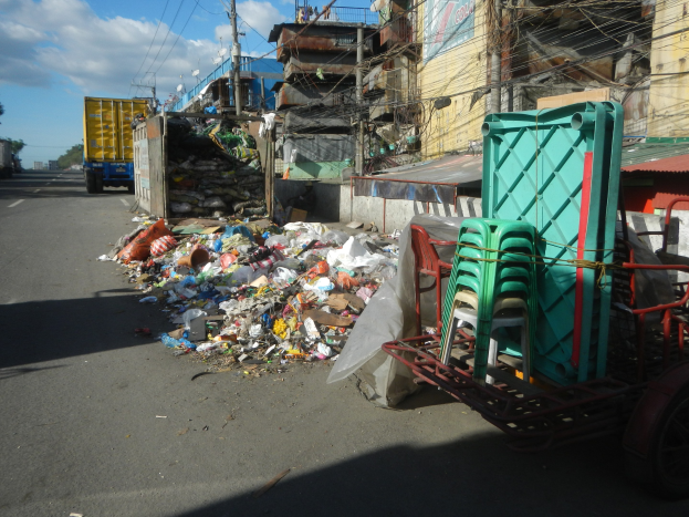Ein Lastwagen neben einem Müllhaufen auf einer Straße, mit einem Wagen mit Plastikstühlen rechts daneben und Gebäuden, Strommasten mit Drähten, Bäumen und einem bewölkten Himmel im Hintergrund.