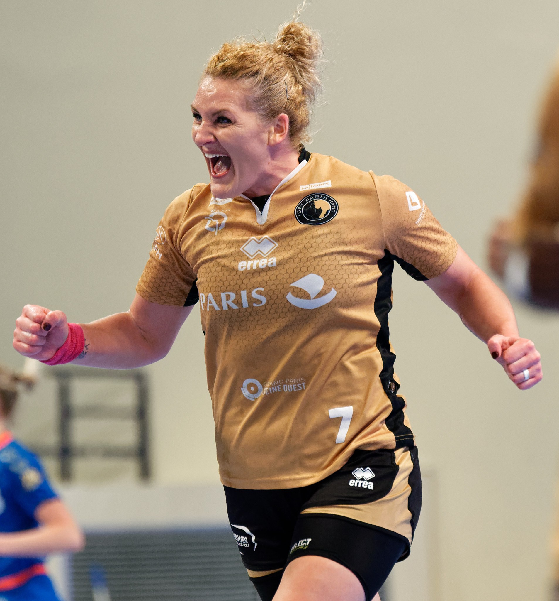 Eine Frau in braunem T-Shirt und schwarzen Shorts, die auf einem Futsalplatz läuft und feiert, mit zwei anderen Frauen im Hintergrund.