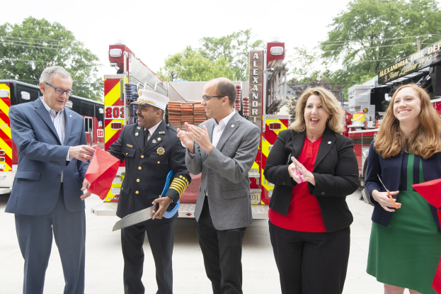 Gruppe von Menschen, die bei einer Ehrung der Alexandria Fire Department klatschen und lächeln, während zwei eine Schere und ein rotes Band halten.