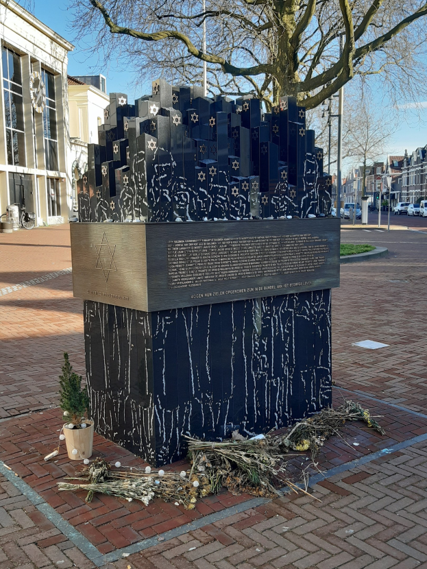 Denkmal für Holocaust-Opfer in einem Stadtplatz mit einem Baum, Topfpflanze, verstreuten Blättern und umgebenden urbanen Elementen.