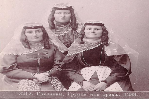 Drei Frauen in traditionellen russischen Hochzeitskleidern mit farbenfroher Stickerei und Perlenbekleidung stehen vor einer einfarbig weißen Wand in einem Schwarz-Weiß-Foto.