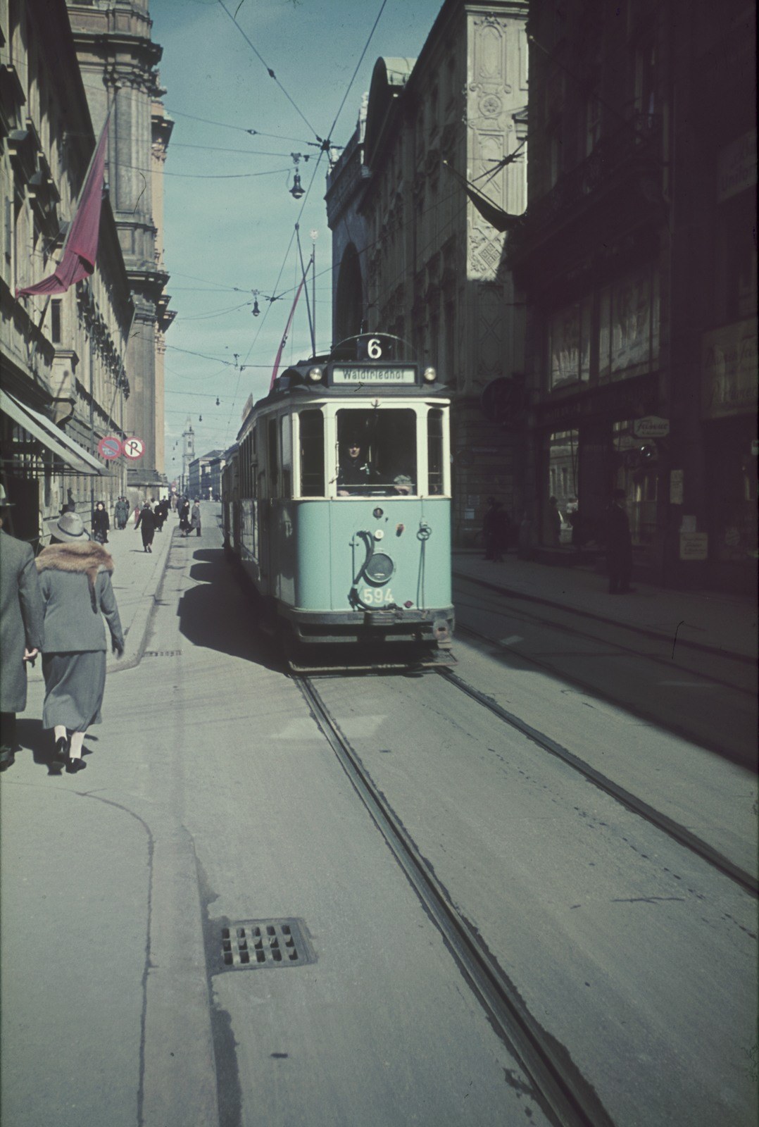 Altes Schwarz-Weiß-Foto einer Straßenbahn, die auf einer Stadtstraße mit Fußgängern auf dem linken Gehweg fährt, mit Gebäuden und Strommasten im Hintergrund unter einem klaren blauen Himmel.