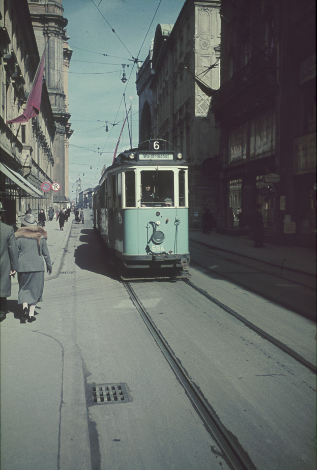 Altes Schwarz-Weiß-Foto einer Straßenbahn, die auf einer Stadtstraße mit Fußgängern auf dem linken Gehweg fährt, mit Gebäuden und Strommasten im Hintergrund unter einem klaren blauen Himmel.