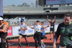 Gruppe von Läufern auf einer Stadionbahn mit Bannern, einem Display, einem Gebäude, Bäumen und einer bewölkten Himmel; einige Läufer tragen Turnschuhe und Hüte.