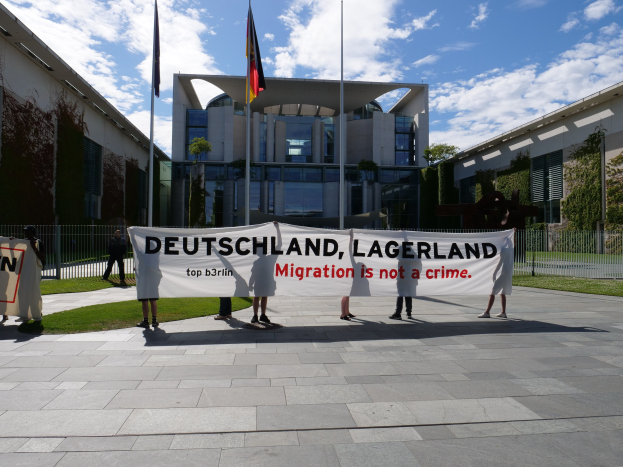 Eine Gruppe von Menschen mit einem Transparent, auf dem "Deutschland, Lagerland Migration ist kein Verbrechen" steht, vor einem Gebäude mit Fenstern, umgeben von Gras, einem Metallzaun, Pflanzen, Bäumen und Fahnenstangen unter einem bewölkten Himmel.