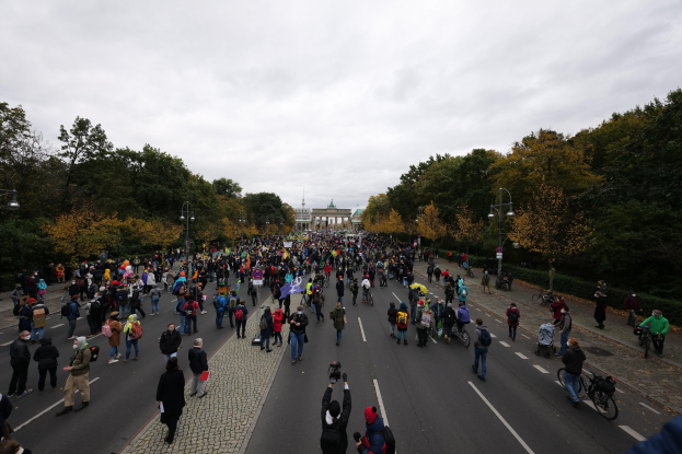 Eine große Gruppe von Menschen marschiert auf einer Demonstration in Berlin durch eine Straße mit Bäumen und Laternen, während einige Kameras halten und ein Gebäude im Hintergrund zu sehen ist.