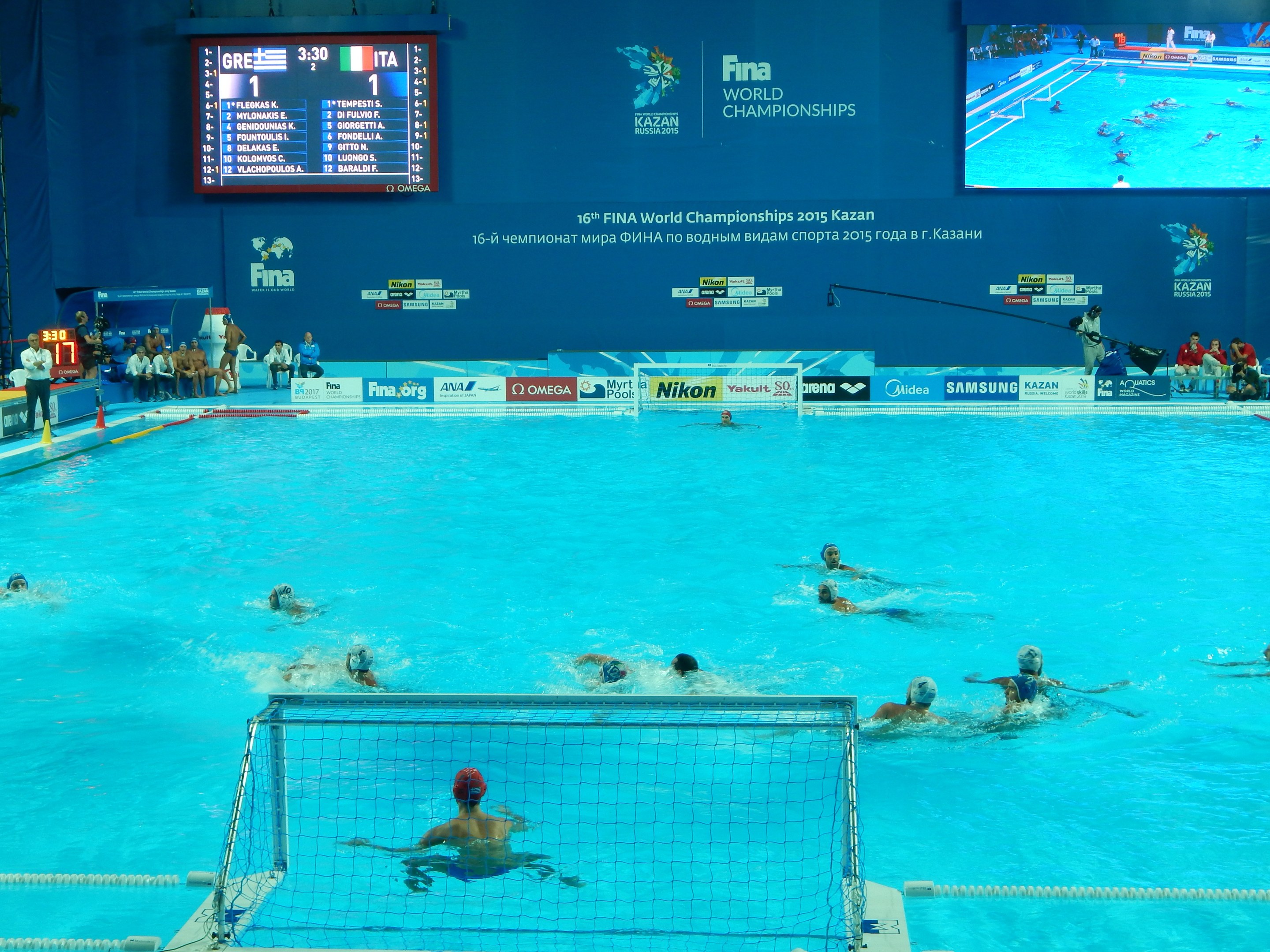Wasserballspieler bei den 2016 FINA-Weltmeisterschaften in Kazan im Pool gegeneinander antretend, mit Zuschauern auf der linken Seite und einer Anzeigetafel im Hintergrund.