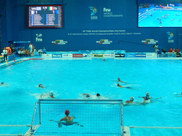 Wasserballspieler bei den 2016 FINA-Weltmeisterschaften in Kazan im Pool gegeneinander antretend, mit Zuschauern auf der linken Seite und einer Anzeigetafel im Hintergrund.