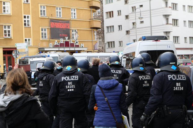 Eine Gruppe von Polizisten steht vor einer Menge von Menschen mit Helmen und Jacken während einer Demonstration in Berlin, Deutschland, mit Fahrzeugen, Gebäuden, Laternenmasten und einem Banner mit Text im Hintergrund und einer Person mit einer Kamera.