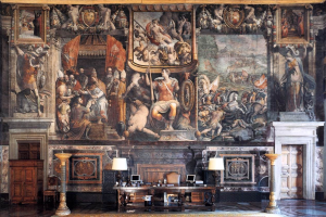 Ein geräumiger Raum im Palazzo della Signoria in Florenz mit einem großen Gemälde, einem Tisch mit Gegenständen, zwei Lampen, zwei Türen und einem Teppich.