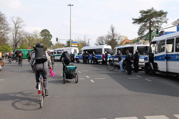 Eine Gruppe von Menschen, die auf Fahrrädern eine Straße entlangfahren, mit Polizeiwagen und anderen Fahrzeugen, einige tragen Helme und haben Taschen oder schieben Kinderwagen, vor dem Hintergrund von Bäumen, Gebäuden und Verkehrsinfrastruktur.