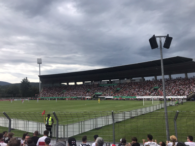 Eine große Menschenmenge schaut einem Fußballspiel in einem bewölkten, eingezäunten Stadion mit beleuchteten Flutlichtmasten zu, umgeben von Bäumen und Hügeln.