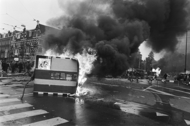 Schwarzes und weißes Foto eines brennenden Busses am Straßenrand mit Menschen in der Nähe, umgeben von Pfählen, einem Schild, Bäumen, Gebäuden und einem bewölkten Himmel.