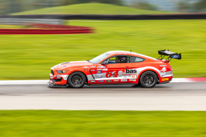 Ein Ford Mustang GT Rennauto, verziert mit Text und Zahlen, fährt auf einer Rennstrecke mit einem Helmträger im Sitz, der Hintergrund ist unscharf, um die Bewegung zu betonen.