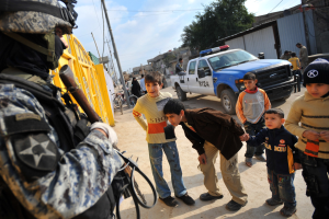 Eine Gruppe von Kindern steht vor einem Polizisten mit einer Waffe, mit Fahrzeugen, Menschen, Fahrrädern und Bäumen im Hintergrund unter einem bewölkten Himmel.