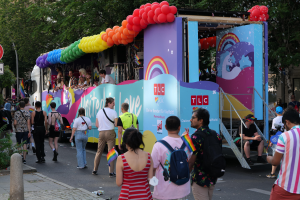 Eine Gruppe von Menschen, die eine Straße entlanggehen, neben einem Lastwagen mit bunten Luftballons, mit Schildern an Pfählen entlang der Straße und Bäumen und Gebäuden im Hintergrund, was auf eine Pride-Parade in Paris hindeutet.