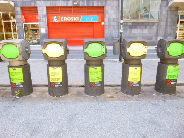 Reihe von Parkautomaten entlang einer Straße mit einem Gebäude mit Glasfenstern und einem Rollladen mit Text im Hintergrund und einem näheren Müllcontainer.