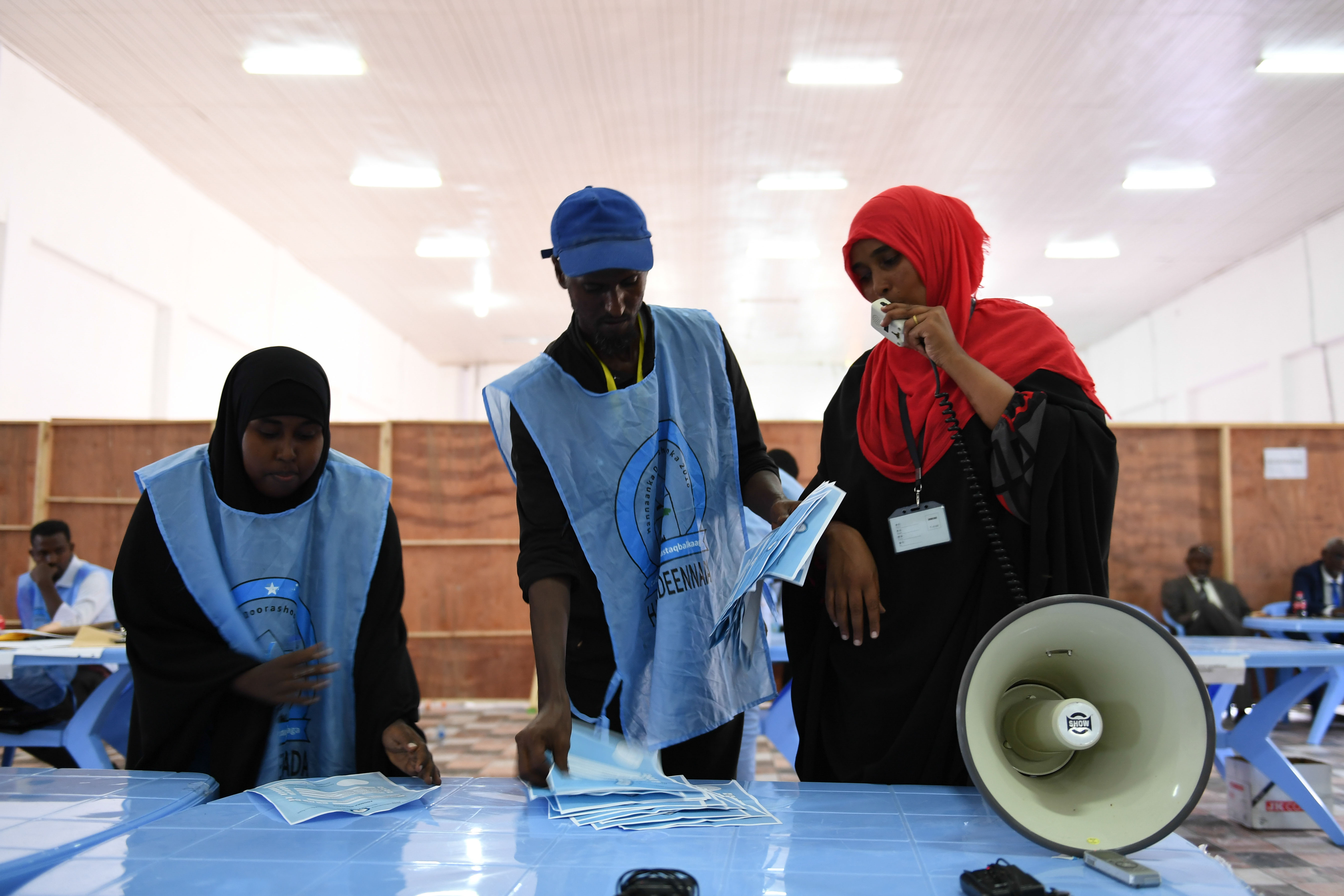 Eine Gruppe von Menschen steht um einen Tisch mit einem Megafon, Papieren und anderen Gegenständen, während andere im Hintergrund auf Stühlen oder stehen unter einer Decke mit Lichtern, in einem, was wie eine Wahllokal aussieht.