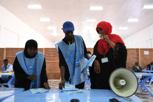 Eine Gruppe von Menschen steht um einen Tisch mit einem Megafon, Papieren und anderen Gegenständen, während andere im Hintergrund auf Stühlen oder stehen unter einer Decke mit Lichtern, in einem, was wie eine Wahllokal aussieht.