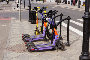 Eine Reihe von E-Scootern, die entlang einer Straße geparkt sind, mit Fußgängern und Fahrradfahrern auf dem Gehweg, einem Müllcontainer im Vordergrund und Gebäuden, Bäumen und Pflanzen im Hintergrund.