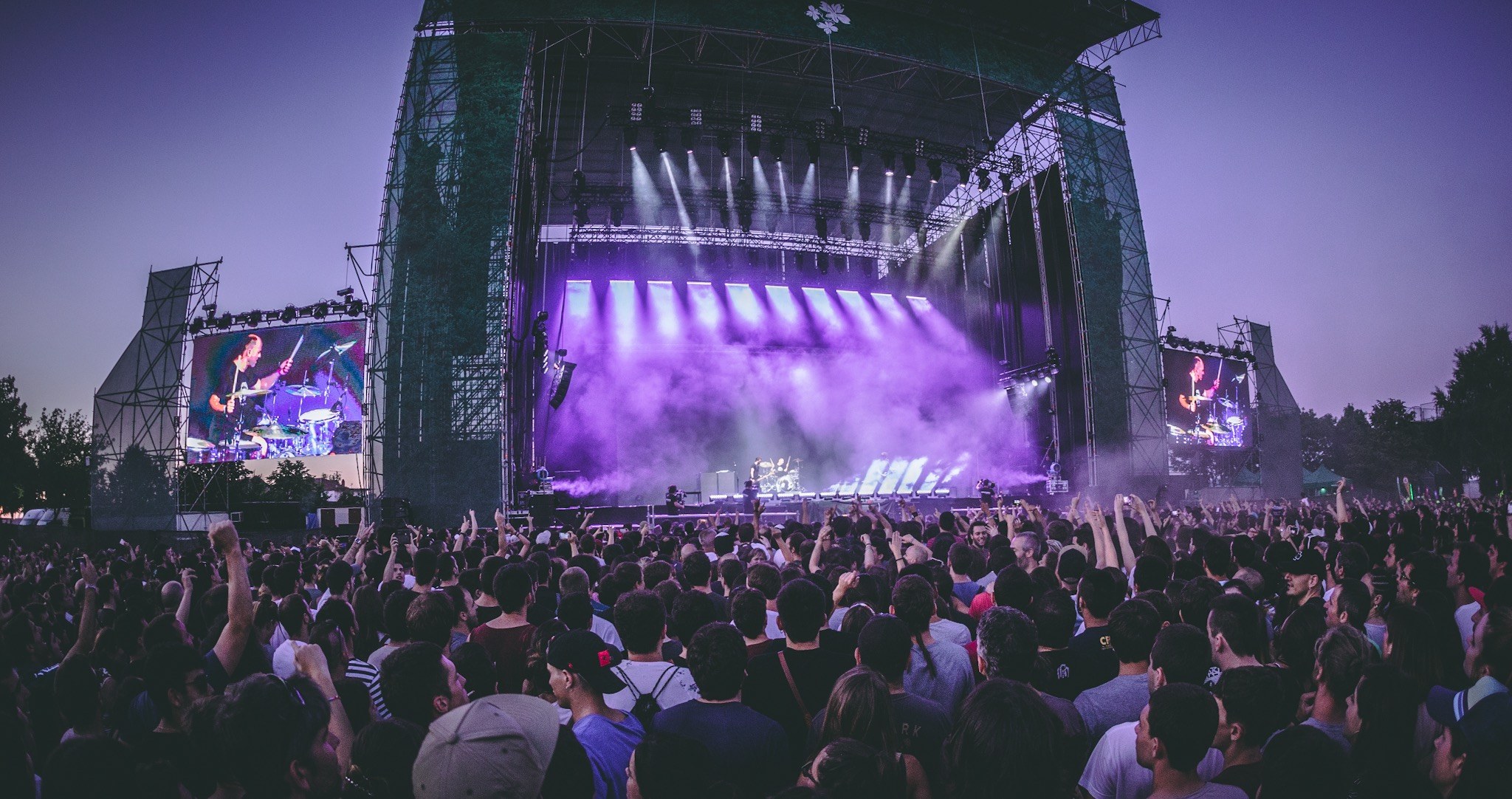 Eine große Menschenmenge bei einem Musikfestival schaut Künstlern auf einer hell erleuchteten Bühne mit Instrumenten und Bildschirmen zu, die vor einem Gebäude, Bäumen und einem klaren blauen Himmel aufgebaut sind.