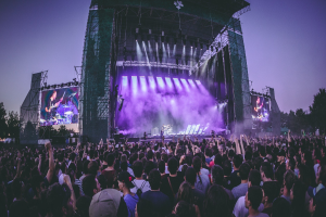 Eine große Menschenmenge bei einem Musikfestival schaut Künstlern auf einer hell erleuchteten Bühne mit Instrumenten und Bildschirmen zu, die vor einem Gebäude, Bäumen und einem klaren blauen Himmel aufgebaut sind.