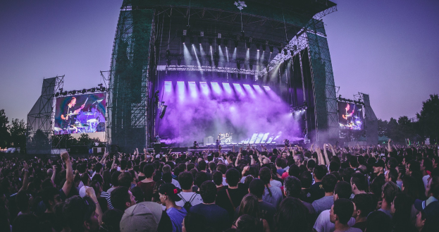 Eine große Menschenmenge bei einem Musikfestival schaut Künstlern auf einer hell erleuchteten Bühne mit Instrumenten und Bildschirmen zu, die vor einem Gebäude, Bäumen und einem klaren blauen Himmel aufgebaut sind.