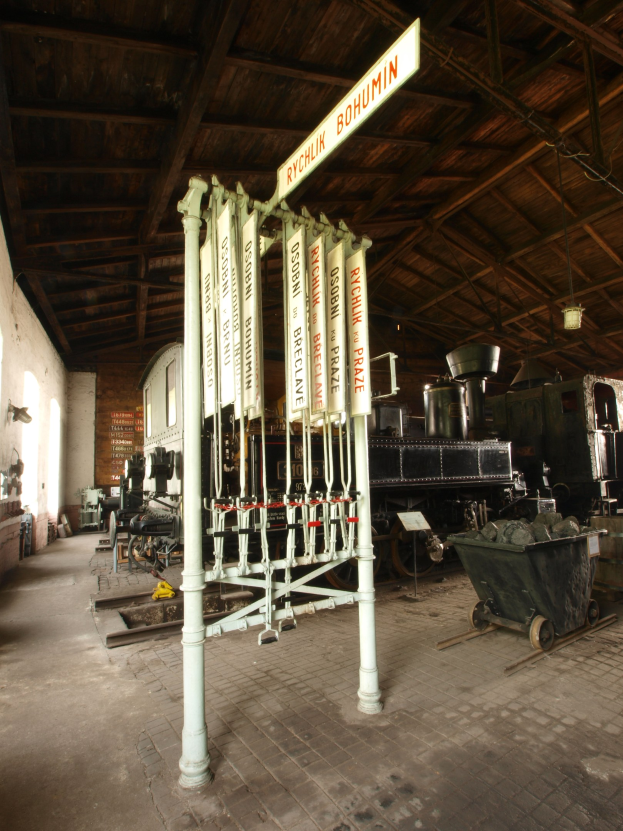 Ein Bahnhof mit einem Zug auf den Gleisen, einem Rollwagen auf dem Boden, einer Signalbox in der Mitte, einem Pfahl mit einer Tafel, einer Wand auf der linken Seite und einer Decke oben.