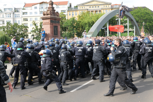 Eine Gruppe von Polizisten in Einsatzausrüstung, einschließlich Helmen, marschiert eine Straße entlang, umgeben von einer Menge, mit Bäumen, Gebäuden, Laternenmasten, einem Bogen, einer Statue auf einem Sockel und einer Fahne mit einem Mast im Hintergrund unter einem bewölkten Himmel.