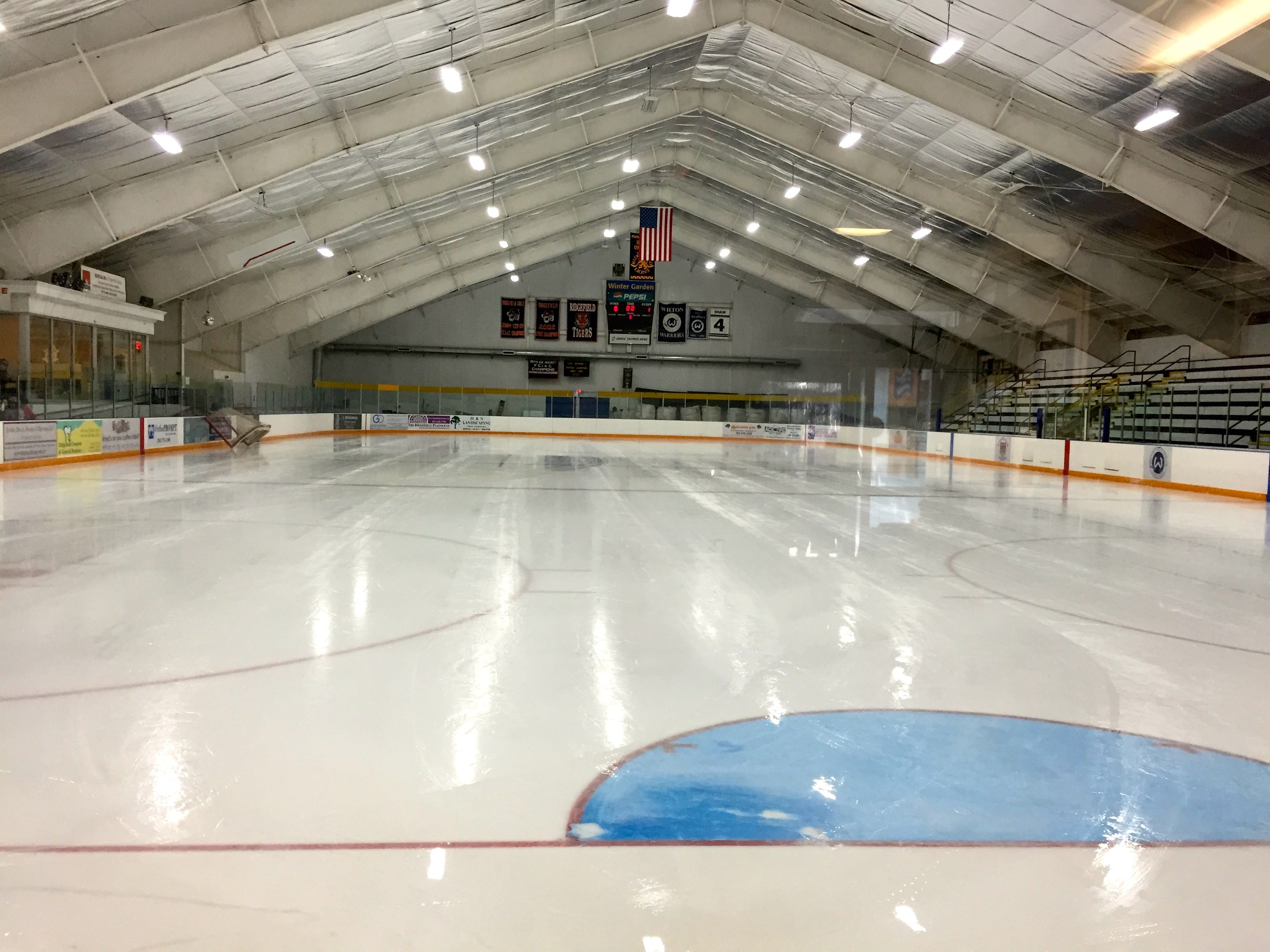 Große Innen-Eisbahn mit Deckenbeleuchtung, Werbetafeln, einer Wand, Treppen mit Geländern und einigen Menschen, was auf eine Sportanlage hinweist, die likely für Hockey verwendet wird.